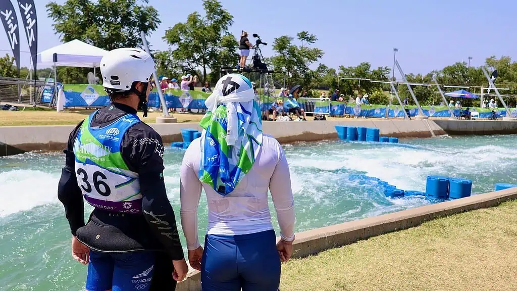 Two athletes wearing helmets stand at the edge of a whitewater slalom course, one with race bib 36 watching the water and spectators.