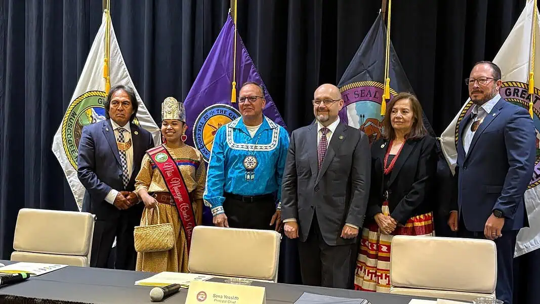 July 2026 ITC Meeting Six people in formal attire stand on a stage with colorful banners and flags behind them, posing for a group photo.