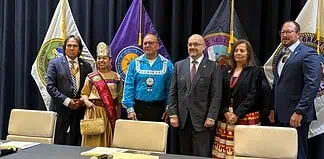 Six people in formal attire stand on a stage with colorful banners and flags behind them, posing for a group photo.