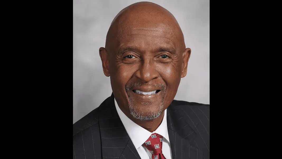MT Berry Professional headshot of a smiling Black man in a dark pinstripe suit and red patterned tie, against a gray backdrop.