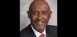 Professional headshot of a smiling Black man in a dark pinstripe suit and red patterned tie, against a gray backdrop.