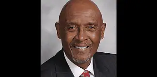 Professional headshot of a smiling Black man in a dark pinstripe suit and red patterned tie, against a gray backdrop.