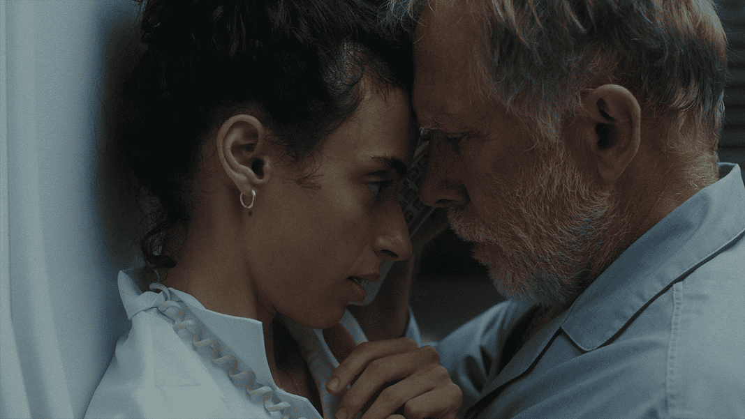 Young woman and an older man press foreheads together in a tender, intimate moment beside a wall.