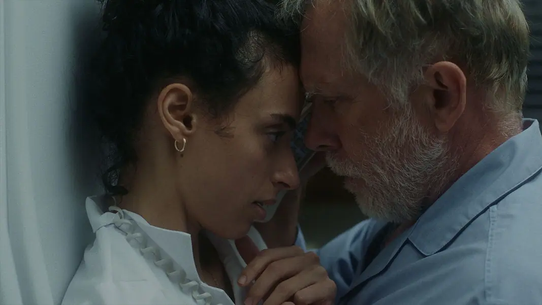 Night Nurse 2 Young woman and an older man press foreheads together in a tender, intimate moment beside a wall.