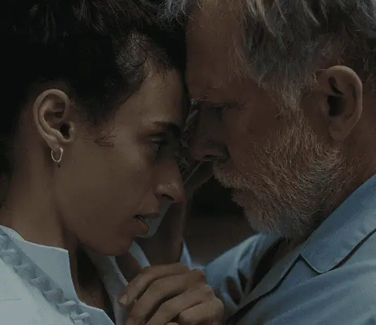 Hypnotic, unsettling ‘Night Nurse’ a piercing portrait of sociopathy Young woman and an older man press foreheads together in a tender, intimate moment beside a wall.
