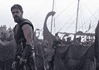 Bearded warrior in dark armor stands at the edge of a tents-filled camp with ships and masts in the misty background.