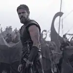 Nolan’s ‘Odyssey’ an epic emotional outburst of present politics, pains Bearded warrior in dark armor stands at the edge of a tents-filled camp with ships and masts in the misty background.
