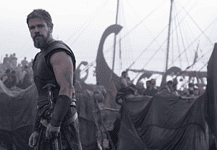 Bearded warrior in dark armor stands at the edge of a tents-filled camp with ships and masts in the misty background.