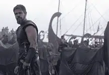 Nolan’s ‘Odyssey’ an epic emotional outburst of present politics, pains Bearded warrior in dark armor stands at the edge of a tents-filled camp with ships and masts in the misty background.