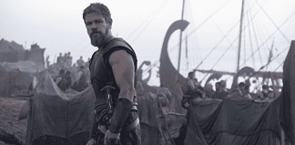 Bearded warrior in dark armor stands at the edge of a tents-filled camp with ships and masts in the misty background.