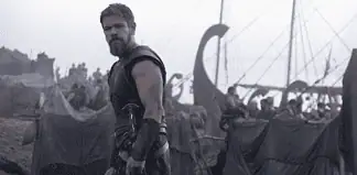 Bearded warrior in dark armor stands at the edge of a tents-filled camp with ships and masts in the misty background.