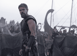 Nolan’s ‘Odyssey’ an epic emotional outburst of present politics, pains Bearded warrior in dark armor stands at the edge of a tents-filled camp with ships and masts in the misty background.