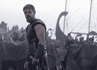 Nolan’s ‘Odyssey’ an epic emotional outburst of present politics, pains Bearded warrior in dark armor stands at the edge of a tents-filled camp with ships and masts in the misty background.