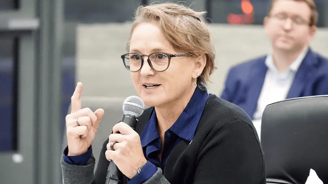 Paula Lewis Woman with glasses speaking into a microphone during a panel discussion in a conference setting.