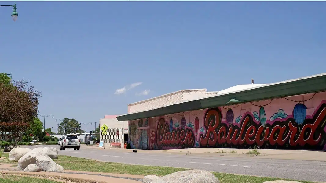 Screenshot Clear blue sky over a street with a long mural spelling 'Boardwalk' along a pink-walled building.