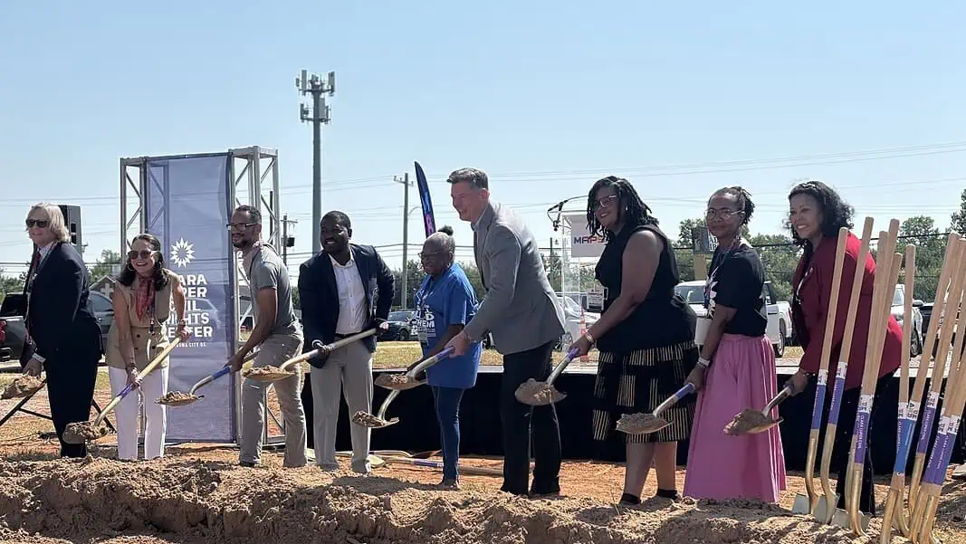 Photo Jul 22 2026, 10 45 46 AM Diverse group at a groundbreaking ceremony, each person with a shovel poised to dig on a dirt mound, banners in background.