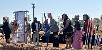 Diverse group at a groundbreaking ceremony, each person with a shovel poised to dig on a dirt mound, banners in background.