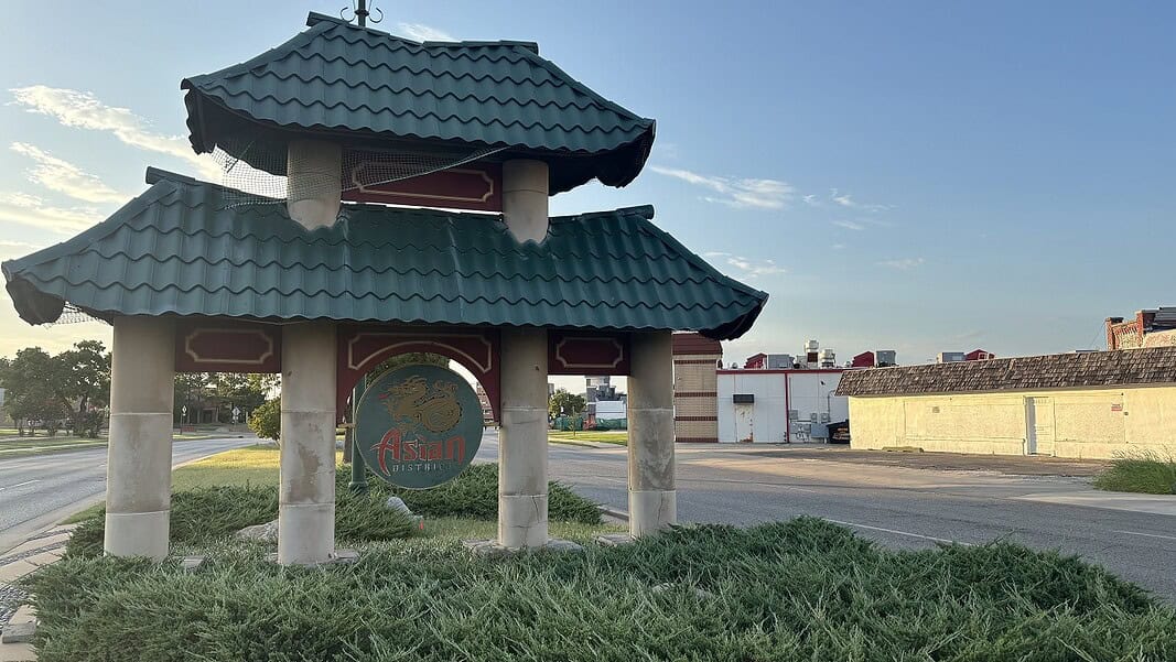 Photo Jul 29 2026, 7 54 03 PM Pagoda-style shelter with a teal, wavy roof over a circular 'Asian' sign on a grassy median by the road.