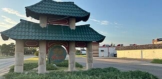 Pagoda-style shelter with a teal, wavy roof over a circular 'Asian' sign on a grassy median by the road.