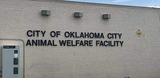 Exterior wall with the sign 'City of Oklahoma City Animal Welfare Facility' and a nearby oxygen warning sign on the right; utility cabinet on the left against a blue sky backdrop.