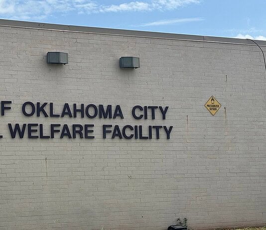 Exterior wall with the sign 'City of Oklahoma City Animal Welfare Facility' and a nearby oxygen warning sign on the right; utility cabinet on the left against a blue sky backdrop.