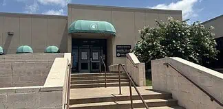 Entrance to a beige building with green curved awnings, concrete stairs, and metal railings under a blue sky.