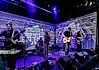 Left-field rockers bring the heat in July music drops Four-piece band performing on a stage with a purple-lit backdrop; singer center, guitarist to the right, keyboardist to the left, drummer in back, with amps and cables visible.