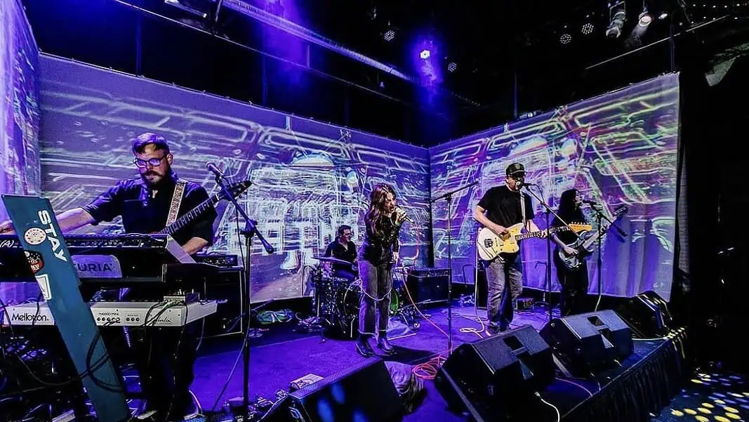 Screenshot Four-piece band performing on a stage with a purple-lit backdrop; singer center, guitarist to the right, keyboardist to the left, drummer in back, with amps and cables visible.