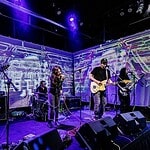 Four-piece band performing on a stage with a purple-lit backdrop; singer center, guitarist to the right, keyboardist to the left, drummer in back, with amps and cables visible.
