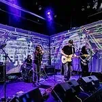 Four-piece band performing on a stage with a purple-lit backdrop; singer center, guitarist to the right, keyboardist to the left, drummer in back, with amps and cables visible.