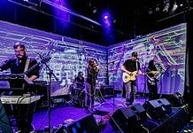 Left-field rockers bring the heat in July music drops Four-piece band performing on a stage with a purple-lit backdrop; singer center, guitarist to the right, keyboardist to the left, drummer in back, with amps and cables visible.