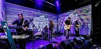 Four-piece band performing on a stage with a purple-lit backdrop; singer center, guitarist to the right, keyboardist to the left, drummer in back, with amps and cables visible.
