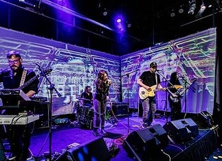 Left-field rockers bring the heat in July music drops Four-piece band performing on a stage with a purple-lit backdrop; singer center, guitarist to the right, keyboardist to the left, drummer in back, with amps and cables visible.