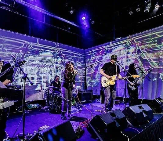 Left-field rockers bring the heat in July music drops Four-piece band performing on a stage with a purple-lit backdrop; singer center, guitarist to the right, keyboardist to the left, drummer in back, with amps and cables visible.