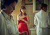 Young woman in a red dress stands at a kitchen island holding a glass, glancing toward the camera while others mingle in the background.