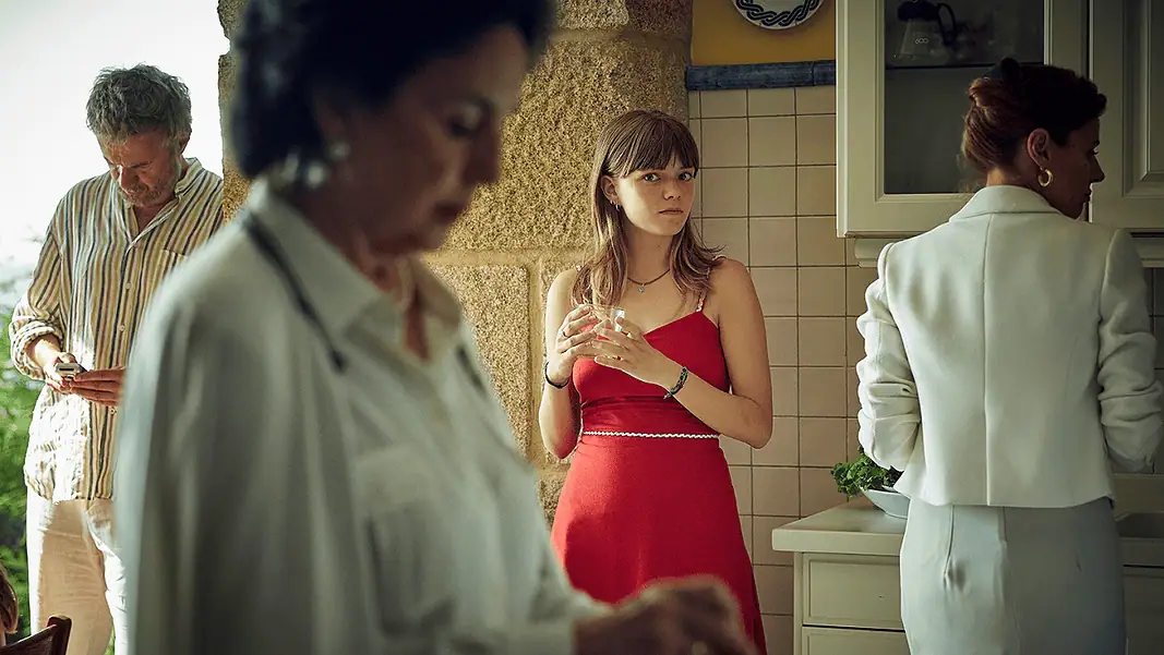 Romeria 1 Young woman in a red dress stands at a kitchen island holding a glass, glancing toward the camera while others mingle in the background.