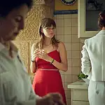 Profound, dreamy ‘Romería’ rewrites the emotional rules of film mystery Young woman in a red dress stands at a kitchen island holding a glass, glancing toward the camera while others mingle in the background.