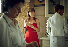 Profound, dreamy ‘Romería’ rewrites the emotional rules of film mystery Young woman in a red dress stands at a kitchen island holding a glass, glancing toward the camera while others mingle in the background.