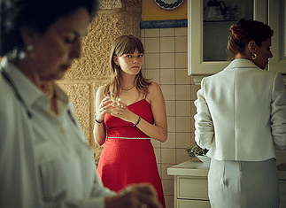 Profound, dreamy ‘Romería’ rewrites the emotional rules of film mystery Young woman in a red dress stands at a kitchen island holding a glass, glancing toward the camera while others mingle in the background.