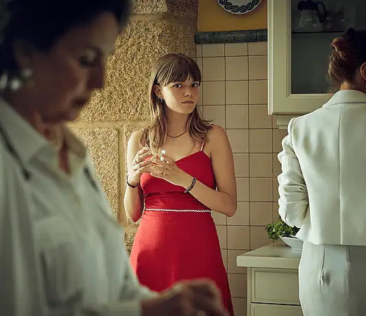 Profound, dreamy ‘Romería’ rewrites the emotional rules of film mystery Young woman in a red dress stands at a kitchen island holding a glass, glancing toward the camera while others mingle in the background.