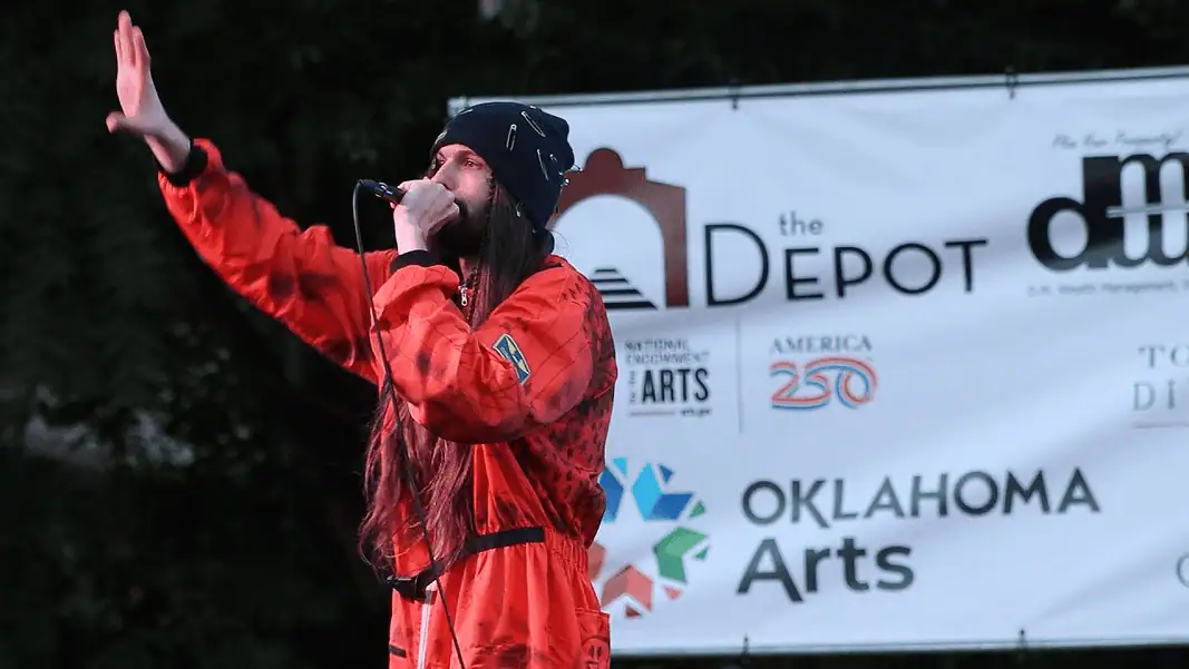 S Reidy live Depot 2 Male performer in a red jumpsuit sings into a microphone on an outdoor stage, with an arts banner in the background.