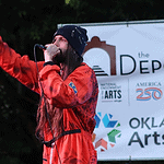 Male performer in a red jumpsuit sings into a microphone on an outdoor stage, with an arts banner in the background.