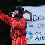 Bring the power of OK’s live music into your home with these new drops Male performer in a red jumpsuit sings into a microphone on an outdoor stage, with an arts banner in the background.