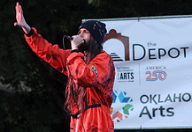 Bring the power of OK’s live music into your home with these new drops Male performer in a red jumpsuit sings into a microphone on an outdoor stage, with an arts banner in the background.