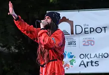 Bring the power of OK’s live music into your home with these new drops Male performer in a red jumpsuit sings into a microphone on an outdoor stage, with an arts banner in the background.