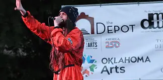 Male performer in a red jumpsuit sings into a microphone on an outdoor stage, with an arts banner in the background.