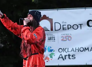 Bring the power of OK’s live music into your home with these new drops Male performer in a red jumpsuit sings into a microphone on an outdoor stage, with an arts banner in the background.