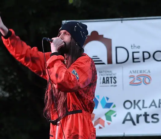 Bring the power of OK’s live music into your home with these new drops Male performer in a red jumpsuit sings into a microphone on an outdoor stage, with an arts banner in the background.