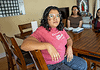 Woman in a pink shirt sits at a wooden dining table, facing the camera, with two others blurred in the background in a home setting.