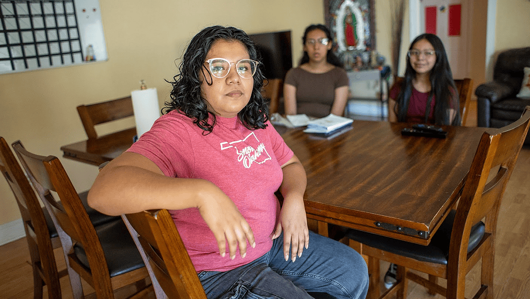 Watch ICE families 1 Woman in a pink shirt sits at a wooden dining table, facing the camera, with two others blurred in the background in a home setting.